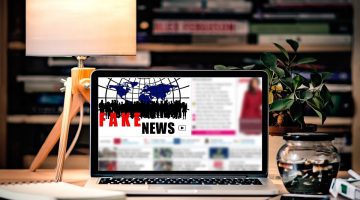Stylized picture of a laptop sitting on a nicely decorated desk displaying the words "fake news" on a blurred out online article