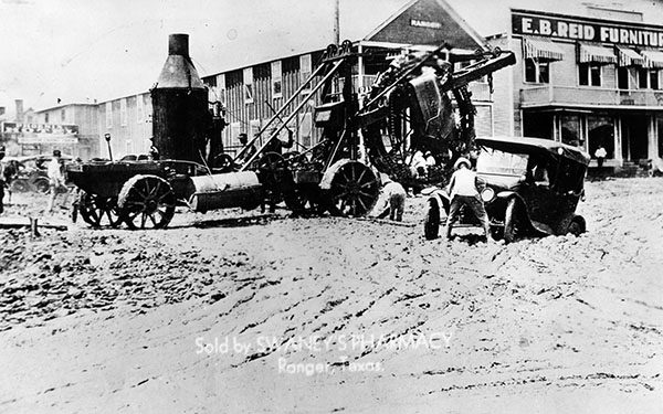 Black and white image of a wagon loaded with oil equipment and a car stuck in a muddy road in a small Texas town