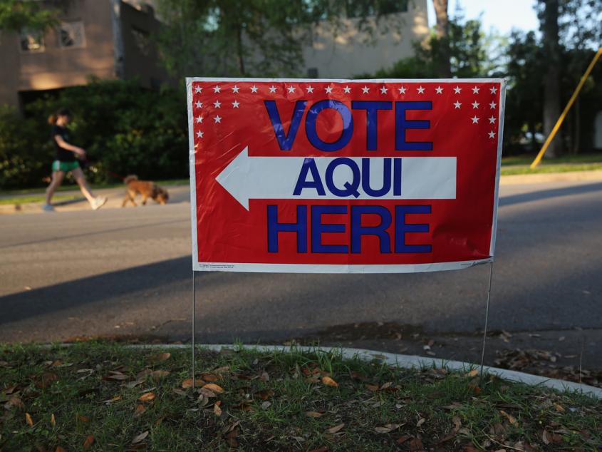 Photograph of a red sign with "Vote Aqui Here" in blue letters with a white arrow