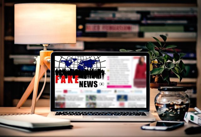 Stylized picture of a laptop sitting on a nicely decorated desk displaying the words "fake news" on a blurred out online article