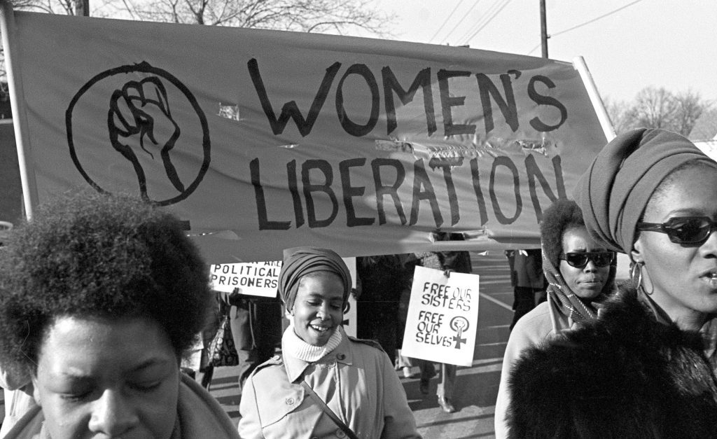 Four black women march beneath a sign reading "women's liberation". In the background, a smaller sign reads: "Free our sisters, free ourselves."