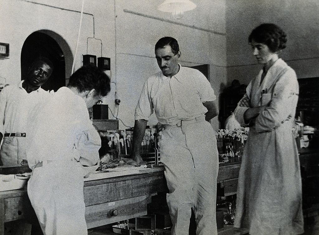 Dr. Hideyo Noguchi (with back to camera) works in a research lab while William Alexander Young and Helen Russell watch.