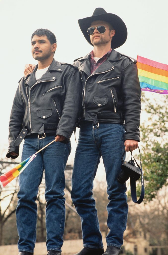 Photograph of two men wearing black leather jackets and jeans at a Gay Pride Event. The man on the left is holding a small rainbow flag and the man on the right is holding a camera and has his arm around the man on the left.