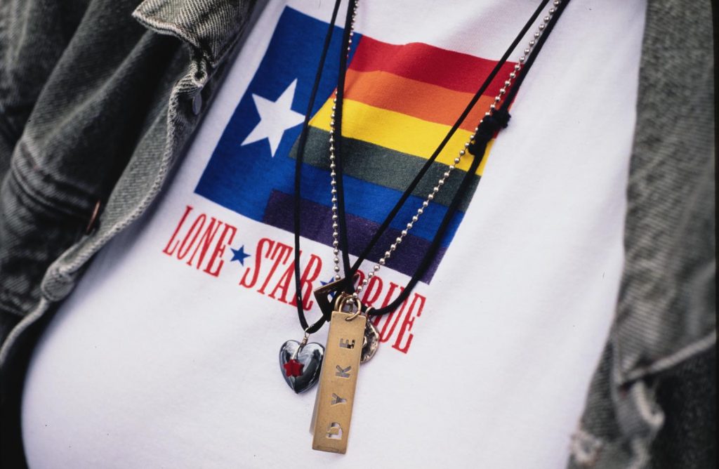 Photograph of a close-up of a white shirt with a rainbow flag on the front that says "Lone Star Pride" underneath and a heart-shaped necklace and rectangular necklace that says "Dyke" laying on top