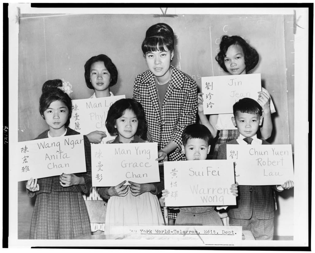 Miss April Lou, a teacher in Manhattan, poses with six Chinese children, recent arrivals from Hong Kong and Formosa, who are holding up placards giving his or her Chinese name (both in ideographs and in transliteration) and the name to be entered upon the official school records.