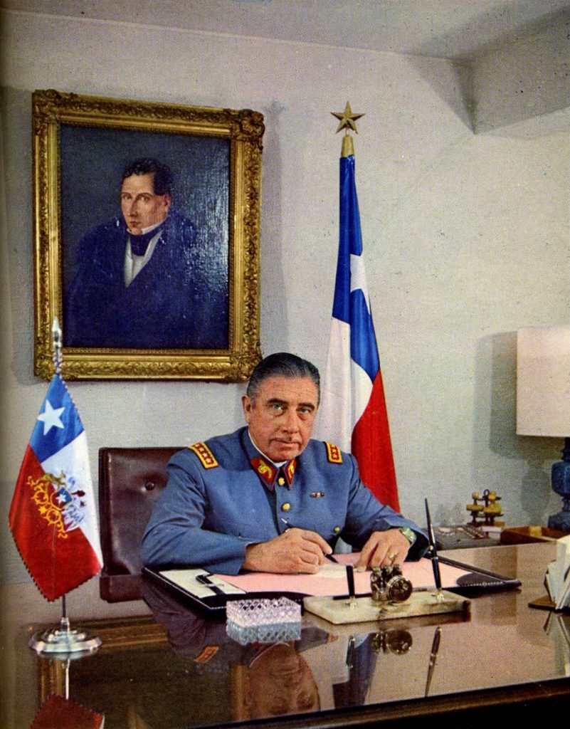Pinochet poses in his office in the attitude of a statesman, pen in hand as if about to sign some important decree. The dictator is flanked by two Chilean flags and behind him is a portrait of Diego Portales, his predecessor as Minister of War and one of the founders of the Republic.