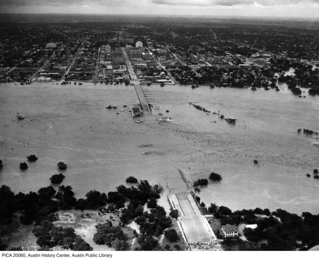 An aerial view of the Colorado River in Austin during a 1930s flood. The Congress Avenue bridge and several city blocks are submerged. A crowd is gathered at the south end of the bridge. The capitol is in the distance. 