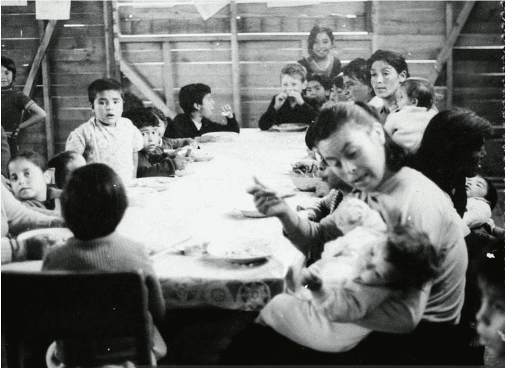 A woman sits at a table while feeding a baby in her lap. The table is surrounded by young children and women.