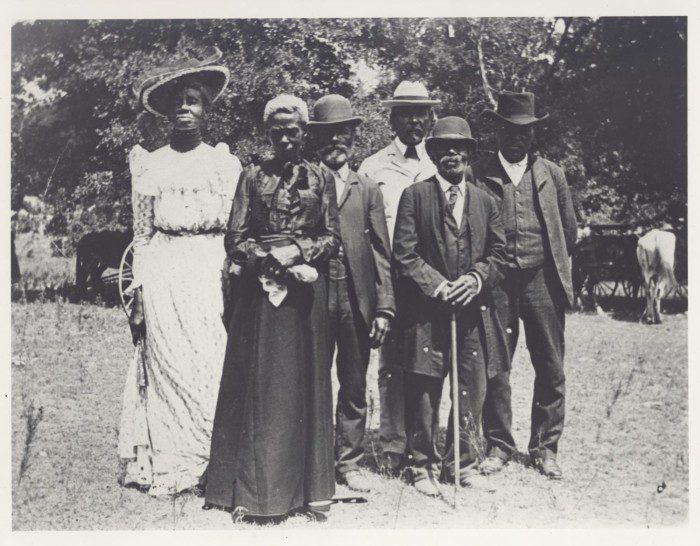 Six Black women and men pose for a photograph outdoors. A woman on the far left wears a floor length white dress and large sunhat, beside her is a woman in a dark dress. The four men wear suits and hats. All look forward toward the camera.