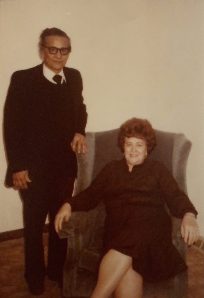 A woman sits in an armchair and smiles. A man stands to her right, resting his arm on the back of the chair. 