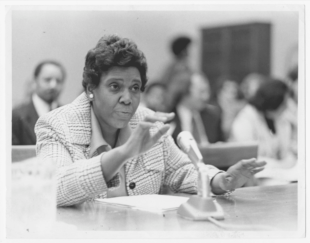 Photograph of Barbara Jordan at a judicial hearing for the House Judiciary Committee. She is wearing a light-colored dress, sitting at a desk with a microphone, and visible from the chest up.