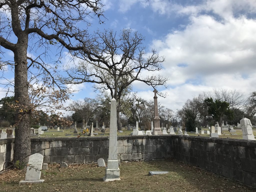 A fall or wintertime photo of Austin's Oakwood Cemetery: under a partly cloudy sky, headstones and obelisks rise amid a cluster of barren trees.