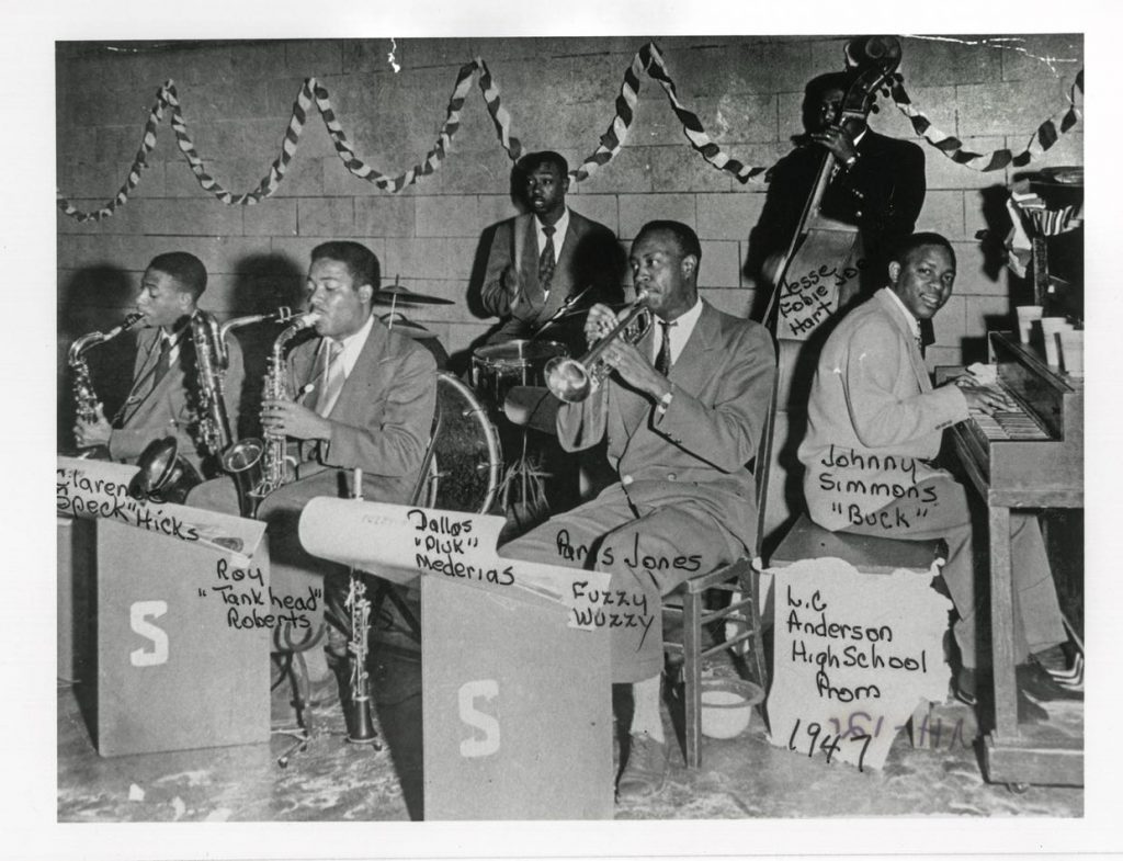 A black-and-white photograph of a band led by musician Johnny Simmons, dated 1947. Handwritten labels identify the six musicians pictured: saxophonists Clarence "Speck" Hicks and Roy "Tankhead" Roberts; drummer Dallas "Pluk" Mederias; trumpeter Paris "Fuzzy Wuzzy" Jones; bass player Jesse Hart, whose nickname is partly obscured; and pianist/bandleader Simmons, nicknamed "Buck." Another label identifies a location, L. C. Anderson High School.