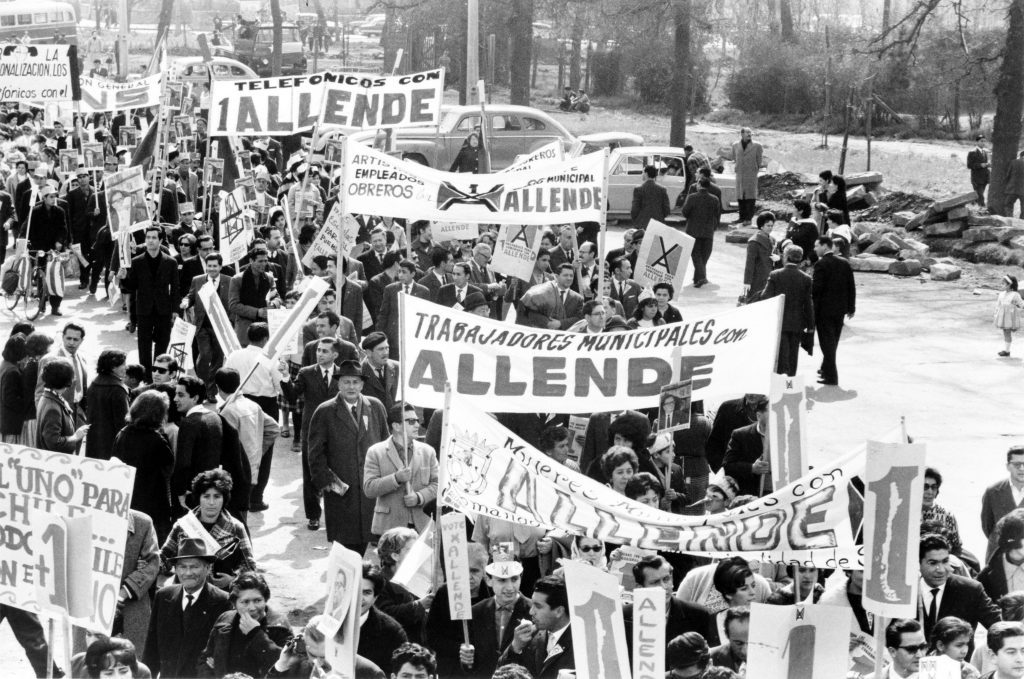 A large crowd marches along a tree-lined street in Santiago in this black-and-white photograph from 1964. Members of the crowd are holding aloft several large banners, all of which indicate support for Salvador Allende. Two banners are easily legible; they read "Telefonicos con 1 Allende" and "Trabajadores municipales con Allende."
