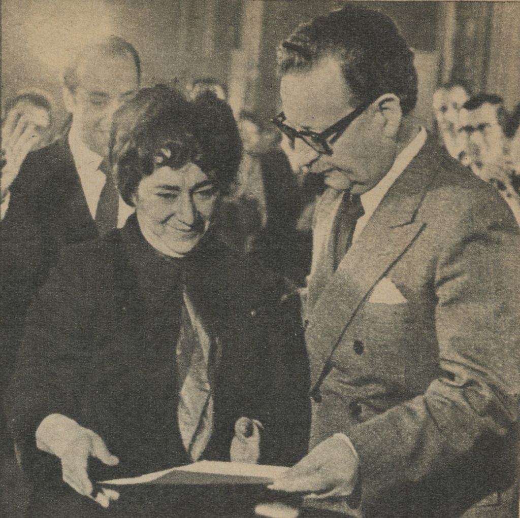 A black-and-white photograph of Salvador Allende and his Minister of Labor and Social Welfare, Mireya Baltra, in the midst of a large crowd of people wearing suits. Both Allende and Baltra are smiling; the President is handing his minister a document.