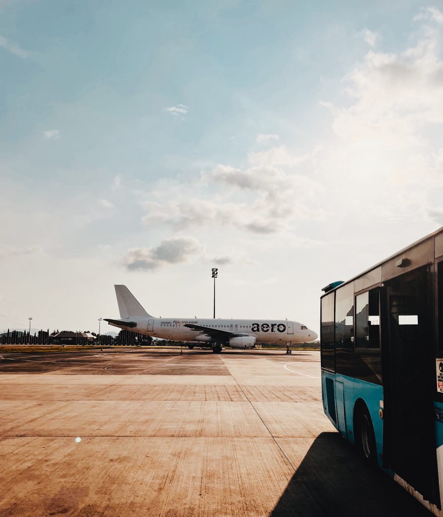 An jet airliner stands on the empty tarmac at an airport in Abuja, Nigeria. The aircraft is painted white and displays the logo of Aero Airlines. In the foreground, a blue low-floored bus is visible.