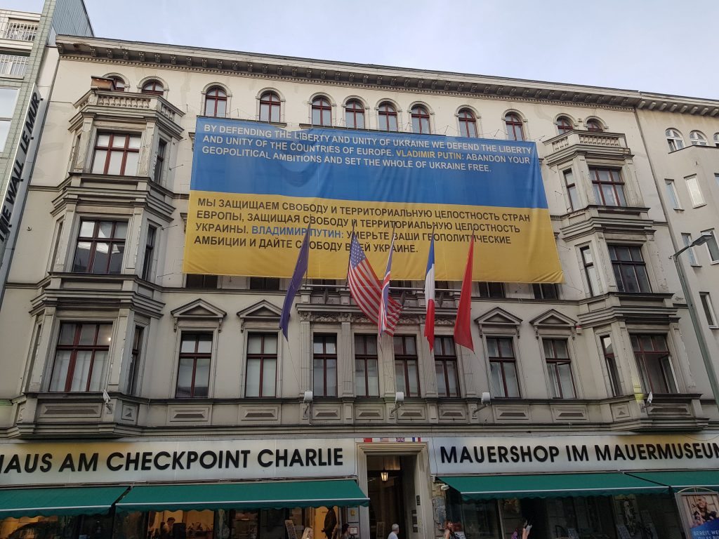 A large banner tinted to resemble a blue-and-yellow Ukrainian flag hangs from the façade of an apartment building in Berlin, Germany, overlooking the "Checkpoint Charlie" museum and giftshop. The banner's text (displayed in both English and Ukrainian) reads: "By defending the liberty of Ukraine we defend the liberty and unity of the countries of Europe. Vladimir Putin: abandon your geopolitical ambitions and set the whole of Ukraine free."