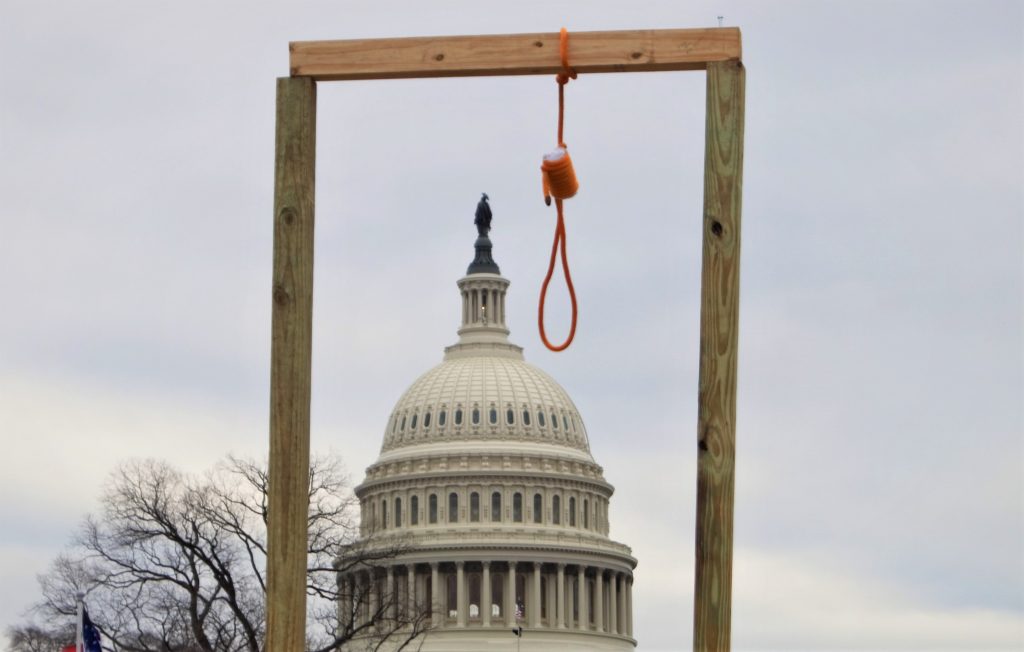 Beneath cloudy skies, a noose hangs from a makeshift gallows erected by rioters during the Insurrection at the U. S. Capitol on January 6th, 2021. The Capitol dome is visible in the background, farmed by the gallows.