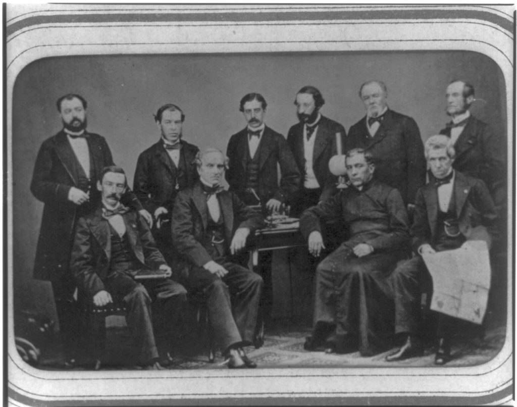 Ten suited men--six standing, four sitting--pose for a photo around a small table. The men are commissioners from Mexico, dispatched to invite Maximilian Habsburg to become that country's Emperor.