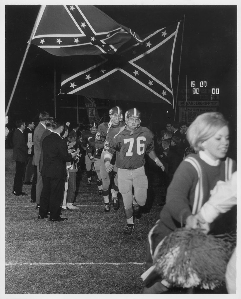 Three football players run onto a playing field applauded by a large group of spectators; a cheerleader leads the charge. Two very large Confederate flags fly overhead.