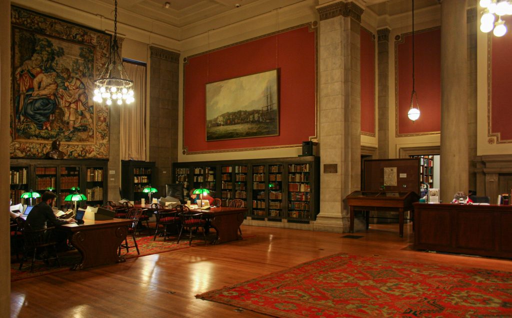 The John Carter Brown Library's MacMillian Reading Room: a large, richly decorated hall with a high ceiling. Low bookshelves and large pieces of art line the walls.; desks with work stations stand in the middle of the room. A few researchers are visible at the desks.