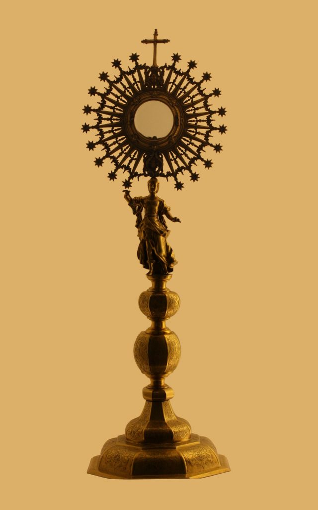 An elaborate ornamental object, golden in color, featuring a tall base, a small figurine, and a large starburst topped by a cross.