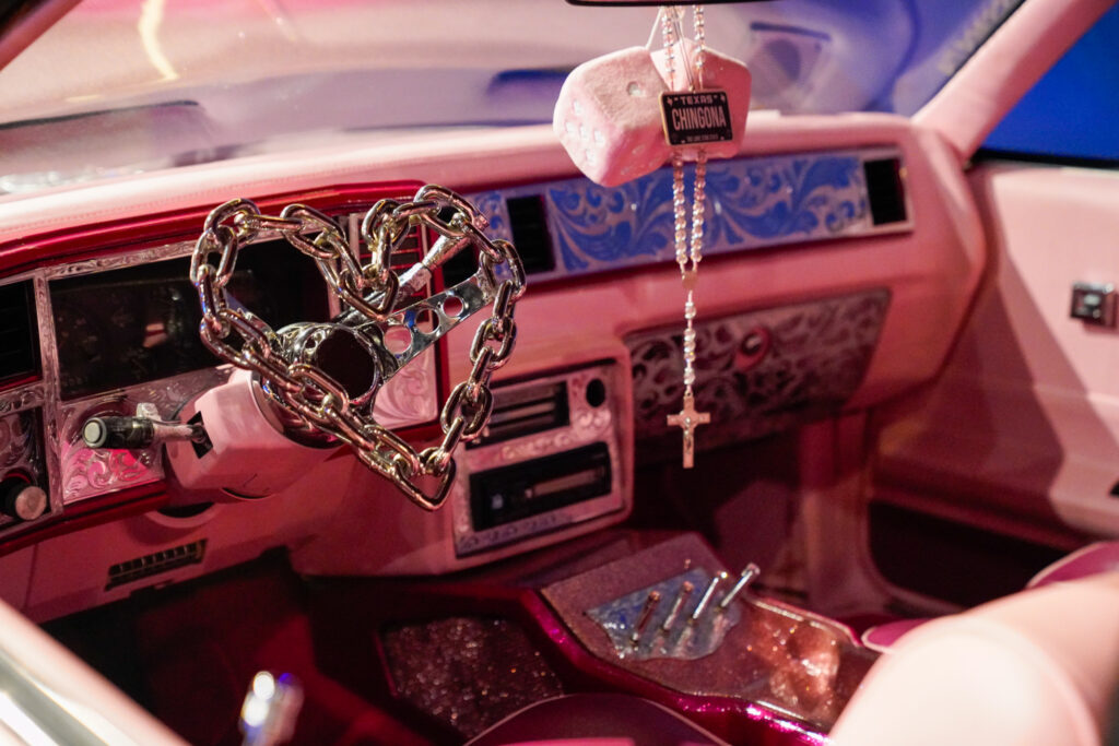 Pink interior of car. Heart shaped wheel is made out of chains. Image from Bullock Museum for Review of Carros y Cultura