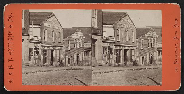 Stereograph showing a man with a rifle sitting outside a commercial building used as a slave market, bearing a sign "Auction & Negro Sales" on Whitehall Street.