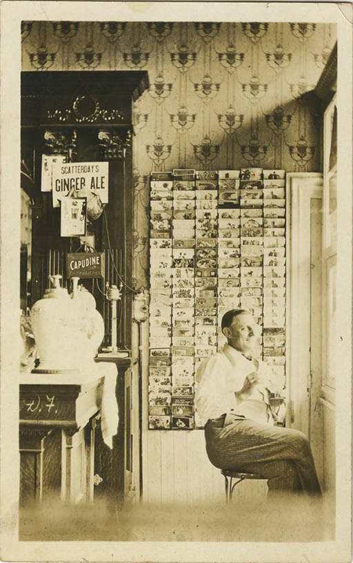 Photo postcard of a man seated in front of a postcard display in a souvenir shop.